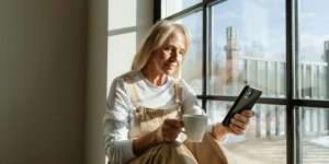 Senior woman sitting by a window with a drink and looking at her phone