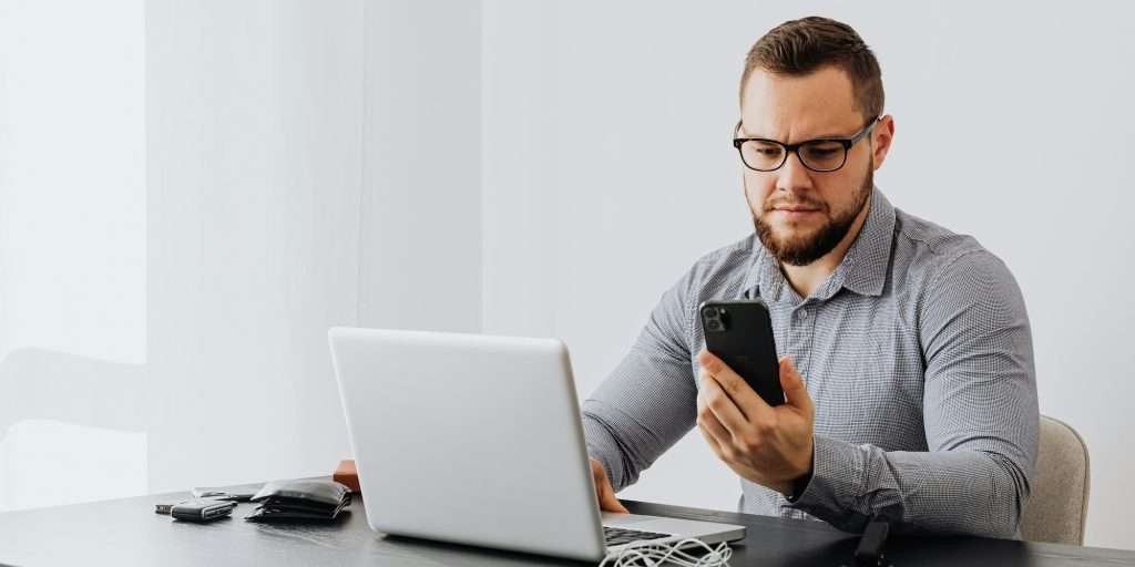 Bearded man in glasses sitting at a desk with a laptop, looking at his phone with a concerned expression.
