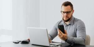 Bearded man in glasses sitting at a desk with a laptop, looking at his phone with a concerned expression.