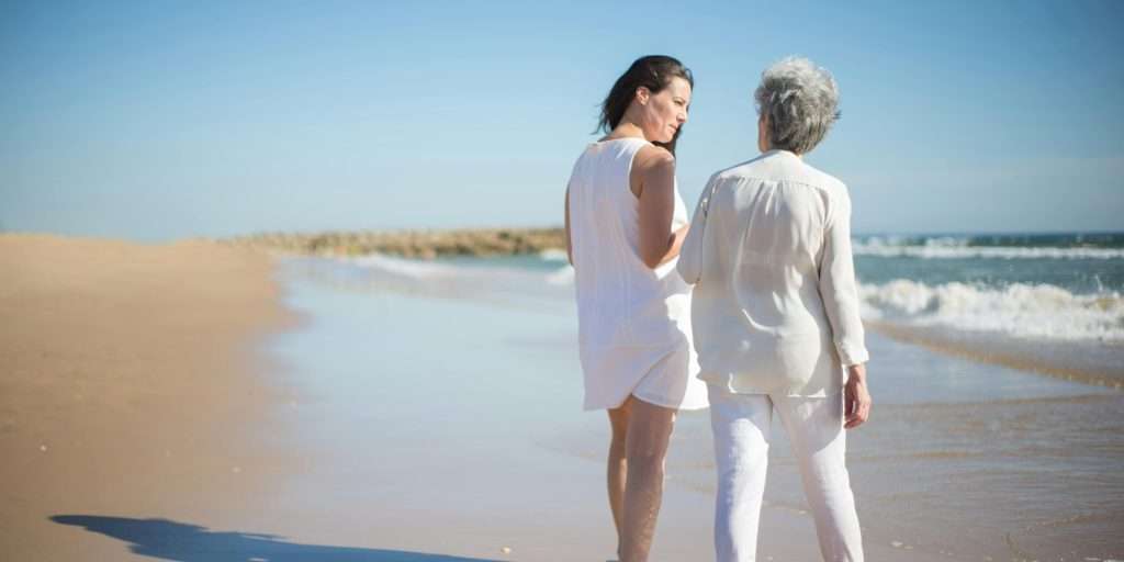 Adult daughter and elderly mother walking together on a beach, viewed from behind